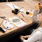 Uczestnicy warsztatów siedzący przy stole z farbami, paletami i szkicami podczas wydarzenia „Jak malujemy swój wizerunek?” organizowanego przez Twój StartUp.