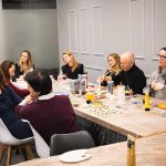 Grupa uczestników siedzących przy dużym stole podczas warsztatów „Jak malujemy swój wizerunek?” organizowanych przez Twój StartUp, z farbami i pracami malarskimi na stole.