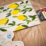 Płótno z namalowanymi cytrynami i liśćmi leżące na stole podczas warsztatów „Jak malujemy swój wizerunek?”, obok palety malarskiej i wizytówki Twój StartUp.