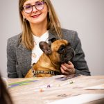 Uczestniczka warsztatów siedząca przy stole z psem na kolanach podczas wydarzenia „Jak malujemy swój wizerunek?” organizowanego przez Twój StartUp.