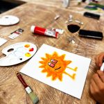 Rysunek uśmiechniętej, pomarańczowej postaci na płótnie obok palety farb, pędzli i kieliszka wina podczas warsztatów „Jak malujemy swój wizerunek?” organizowanych przez Twój StartUp.